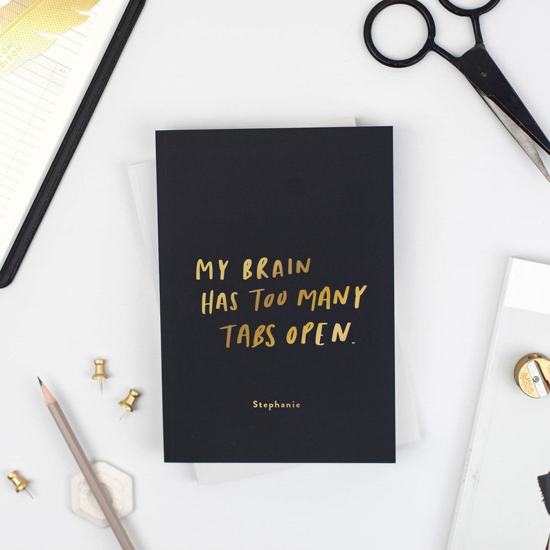 a black coloured A4 notebook with gold text on the front reading 'my brain has too many tabs open' - surrounded by other stationery essentials.