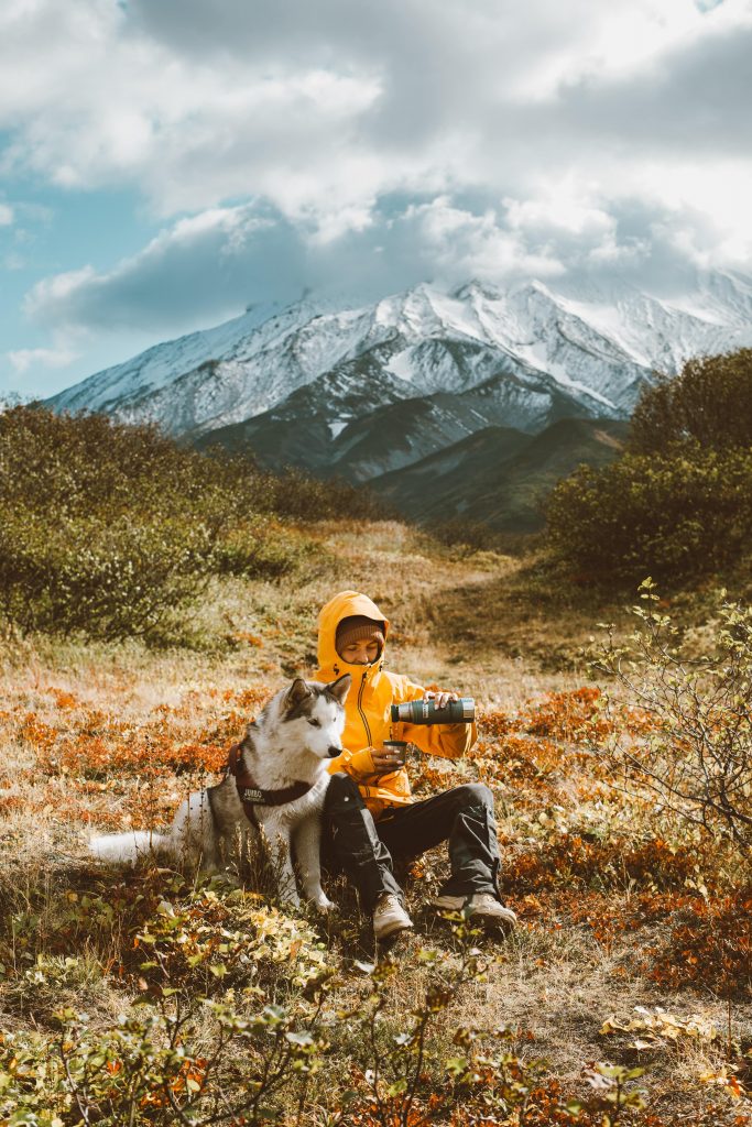 a person pictured sitting upon a large ray of mountains, with their husky, whilst wearing a bright yellow raincoat and holding a camera.