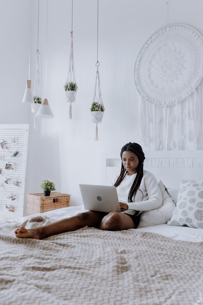 a woman, wearing a white jumper, is sat on her bed with legs outstretched and typing on a laptop that is lay upon her legs.