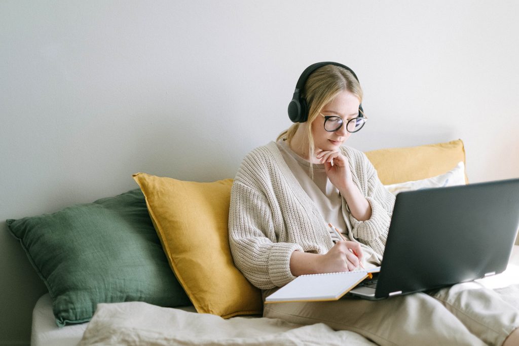 a woman, wearing an ivory jumper, glasses and headphones, is sat on her bed with legs outstretched and typing on a laptop that is lay upon her legs.