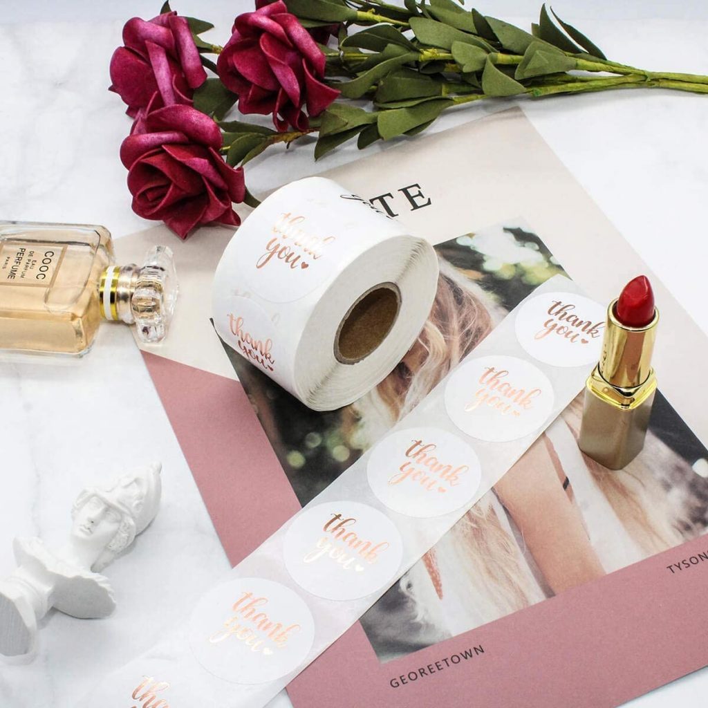 rose gold 'thank you' stickers on a roll, upon a desk with magazines and cosmetics.