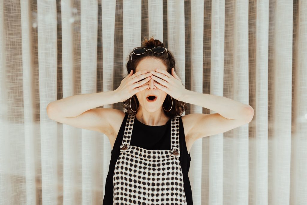 a woman standing in front of vertical blinds, wearing dungarees and a shocked expression whilst covering both eyes with her hands.