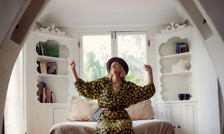 woman wearing a lemon-patterned dress is sat upon a chair with both hands raised in celebration.