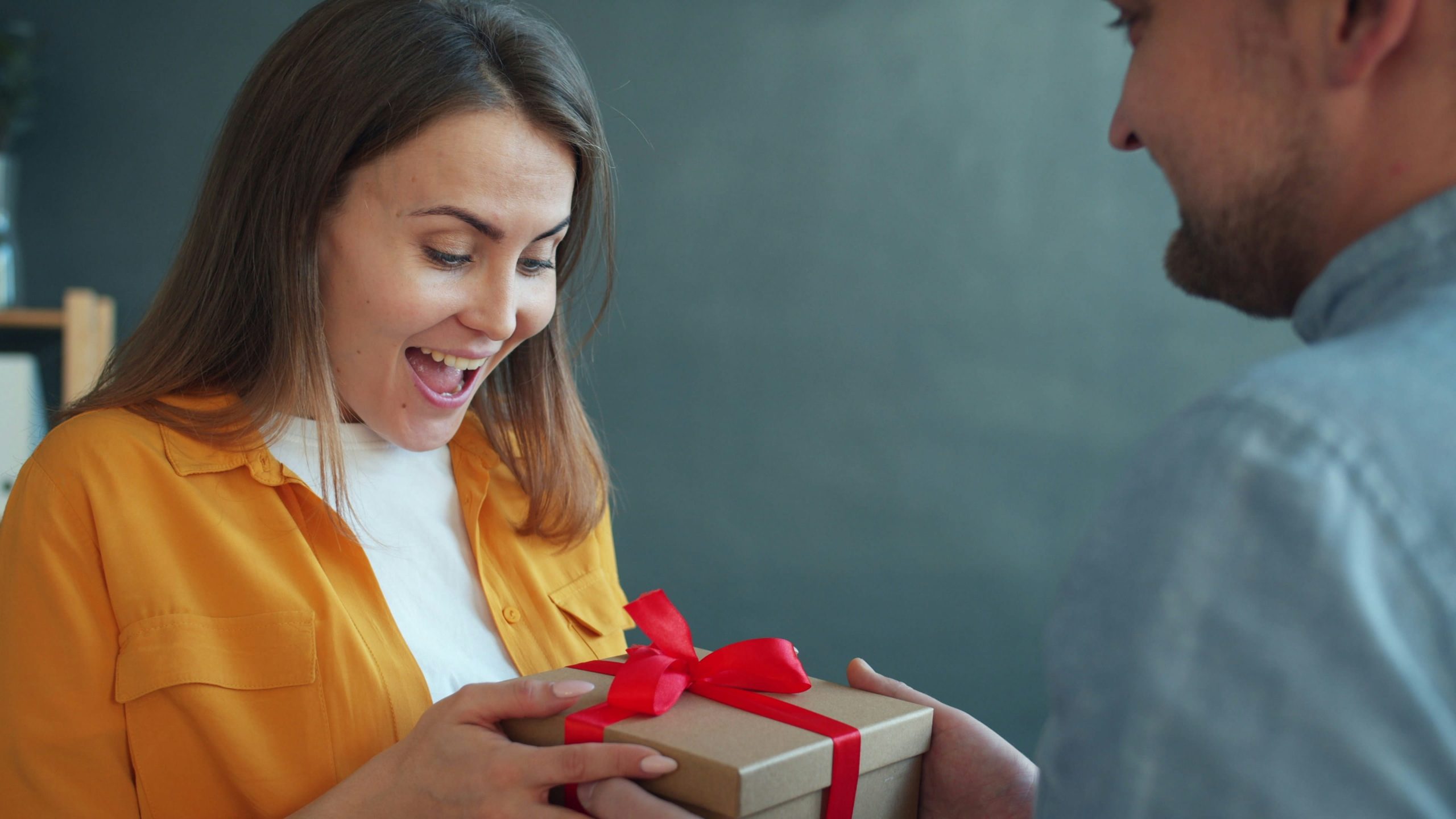 a woman wearing an orange shirt, showing a surprised but happy expression as a man hands her a red ribbon wrapped brown box.