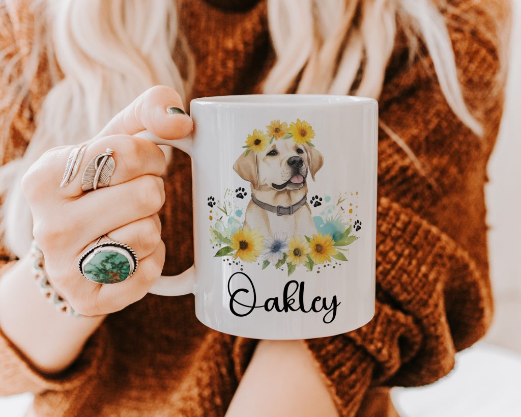 a white ceramic mug being held close by someone with blonde hair, wearing a thick brown jumper. the mug features a watercolour design of a golden / yellow lab wearing a flower crown alongside being surrounded by greenery and more sunflowers - followed by a personalised name.